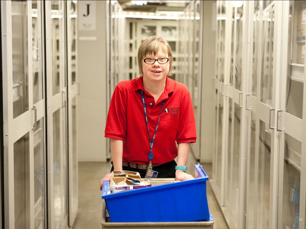 A young woman wheeling a blue box on a cart down a hallway lined with glass doors